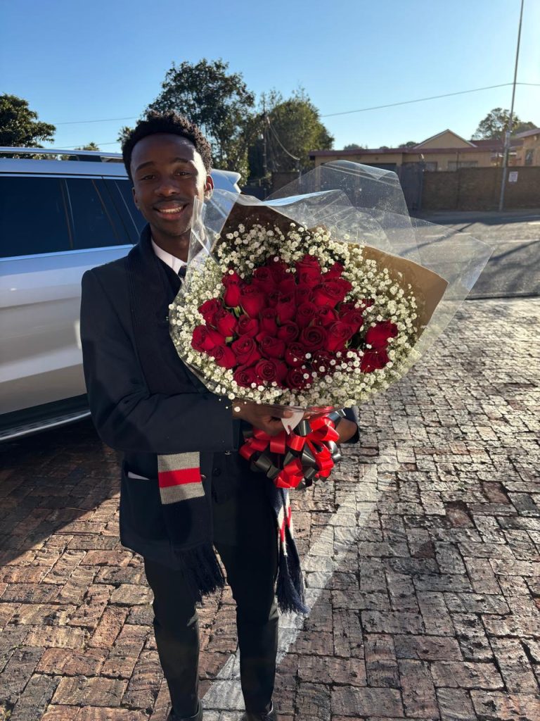 A well-dressed young man smiles warmly while holding a large bouquet of red roses wrapped in baby's breath and cellophane. He stands outdoors on a cobblestone driveway next to a white SUV under clear blue skies. The bouquet is elegantly tied with a black and red ribbon, creating a romantic and stylish presentation—perfect for gifting on special occasions such as anniversaries, Valentine’s Day, or prom. Fresh flowers proudly supplied by Cinnamon Lane Flower Market in Gauteng.